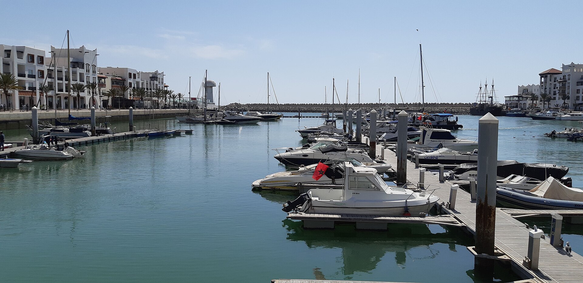 Rows of yachts and fishing boats moored at the Agadir Marina, with whitewashed waterfront buildings and palm trees along the quay, Morocco