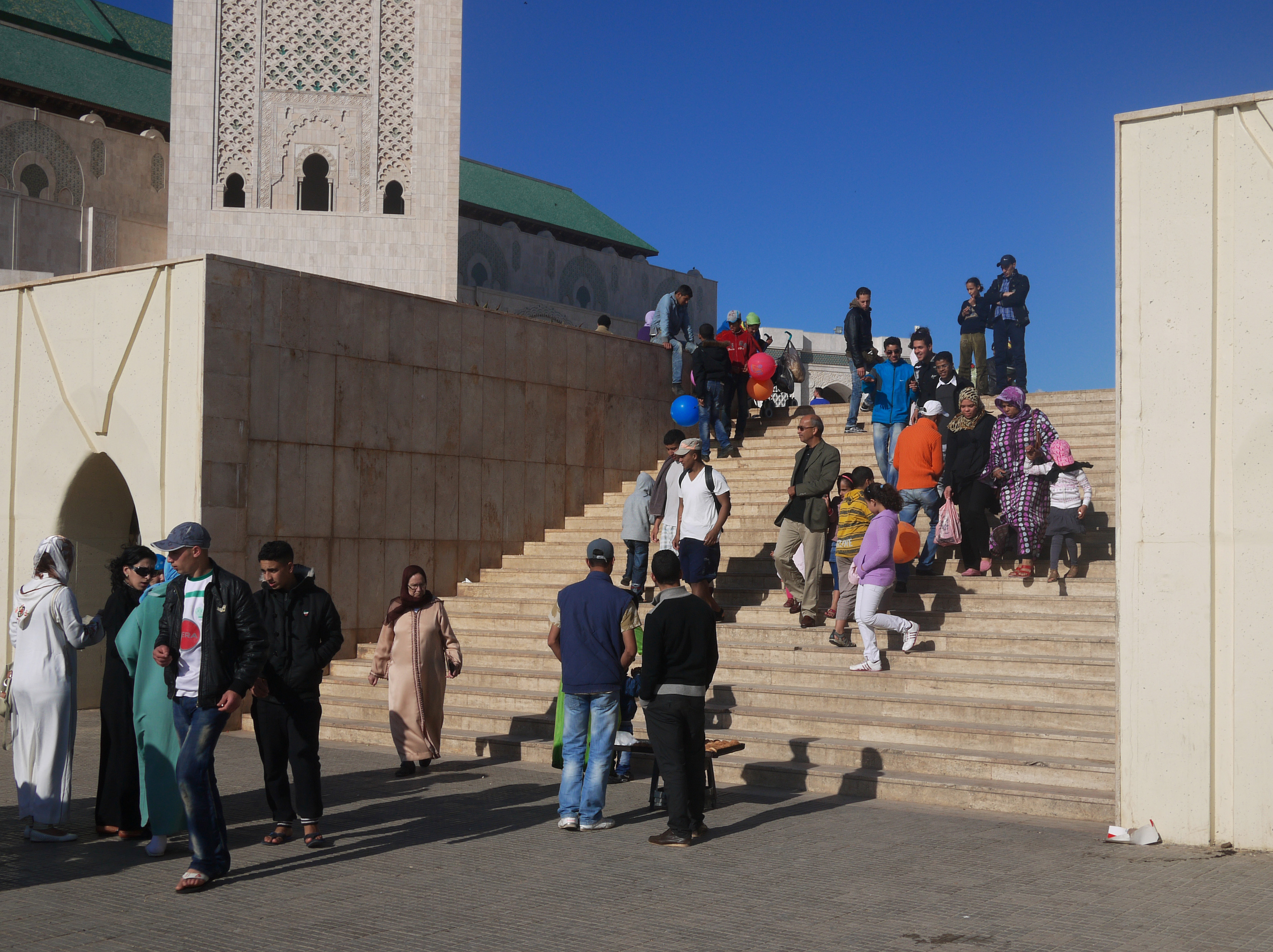 Crowds descending the marble steps beneath the ornate minaret of the Hassan II Mosque in Casablanca, Morocco, under a clear blue sky