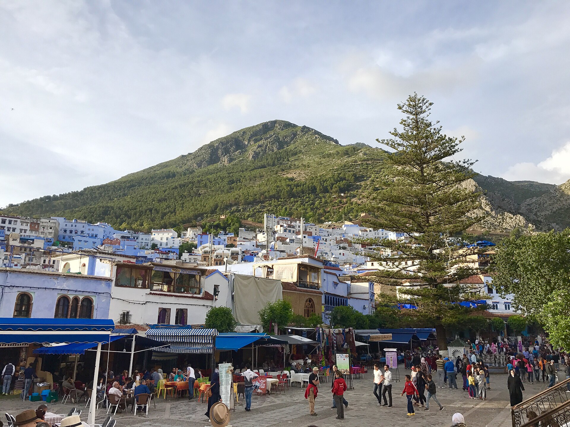The main plaza of Chefchaouen (Uta el-Hammam) full of market stalls and cafe tables with the Rif Mountains rising behind, Morocco