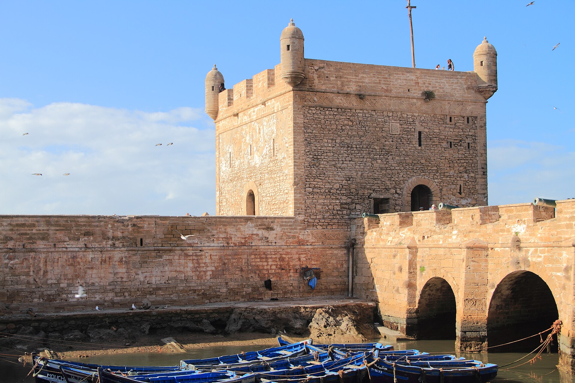 Blue wooden fishing boats moored in the harbor beneath the stone watchtower of the Essaouira citadel, Morocco