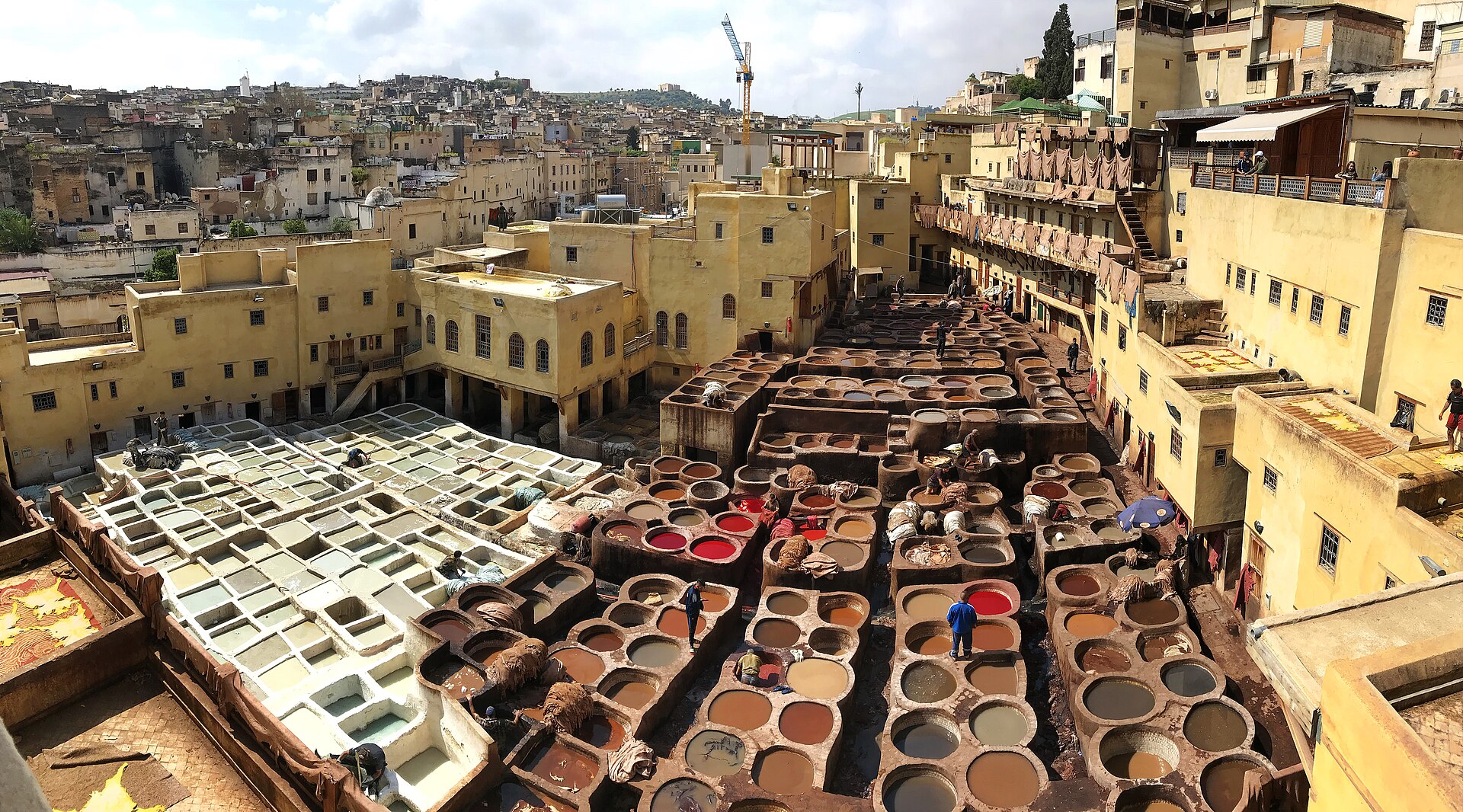 Rows of round stone dye pits filled with colorful natural dyes at the Chouara Tannery in Fes, Morocco, with the medina hillside behind