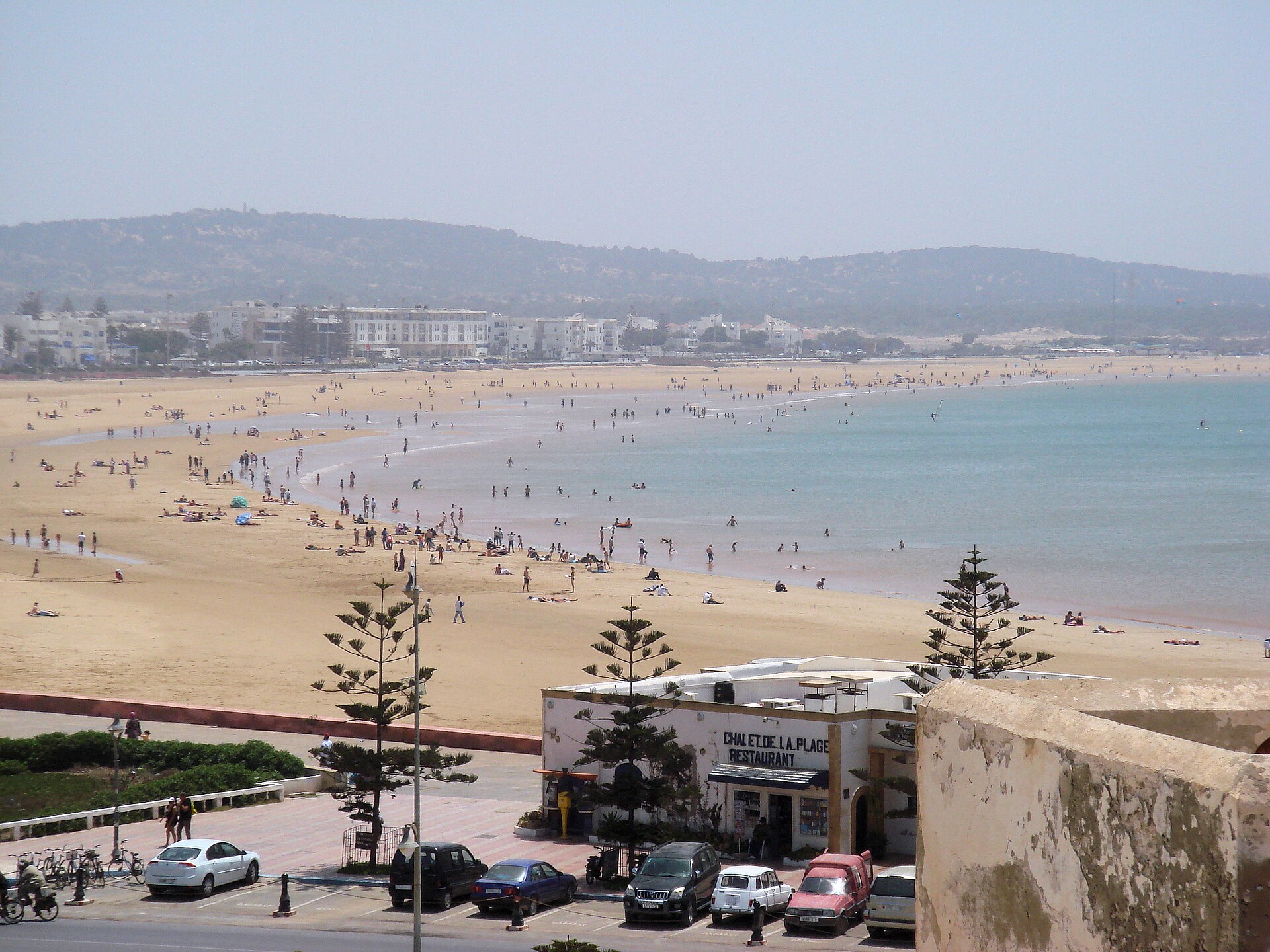 Wide view of Essaouira's sandy beach and Atlantic coastline in Morocco, with the promenade, parked cars, and pine trees in the foreground and hazy coastal hills beyond